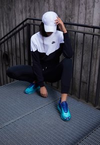 Sportowy zestaw Nike składający się z rozpinanej bluzy w czarno-białe bloki kolorystyczne, czarnych dresów i niebieskich butów sportowych. Na głowie założona biała czapka.