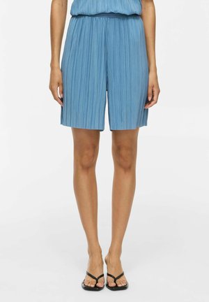 Femme portant un short plissé bleu clair et des sandales noires à bout ouvert, debout sur un fond blanc uni.