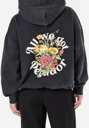 Person iført sort hættetrøje med farverigt blomsterdesign og buet tekst "All We Got Pegador" på ryggen.