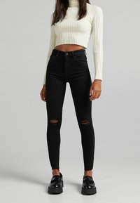 Svarta högmidjade smala jeans med slitna knädetaljer, matchade med en vit rutig kort topp. Slät tyg, åtsittande design och subtil textur.