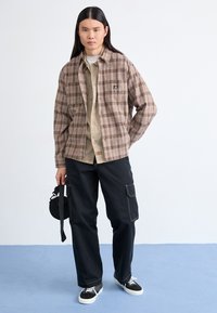 Rutet brun skjortejakke over en beige skjorte med knapper, sammen med svarte cargo-bukser. Svarte sneakers har et teksturert mønster. Sirkulær veske i hånden.
