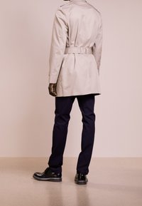Trench coat beige con vita cintata, spalline sulle spalle e maniche lunghe, abbinato a pantaloni slim-fit blu navy e stivali neri con lacci.