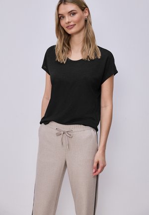 Kobieta w czarnej bluzce z krótkim rękawem i beżowych spodniach z troczkiem oraz bocznymi paskami, stojąca na jednolitym jasnym tle.