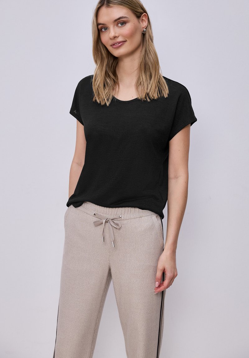 Femme portant un haut noir à manches courtes et un pantalon beige à cordon avec des bandes latérales, debout devant un fond clair uni.