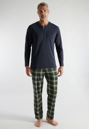 Erwachsener Mann steht barfuß und trägt ein marineblaues Langarm-Henley-Shirt sowie grüne und marineblaue karierte Schlafanzughosen vor weißem Hintergrund.