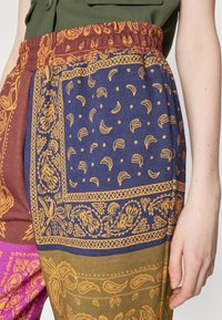 Pantalones de patrones multicolores que presentan una mezcla de tejidos en púrpura, azul marino y verde oliva. El diseño incluye motivos florales y de paisley con acentos en dorado.