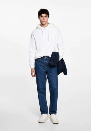 Felpa bianca con cappuccio, jeans blu, sneakers bianche, tiene una giacca blu navy. Abbigliamento informale con vestibilità standard e materiale in cotone.