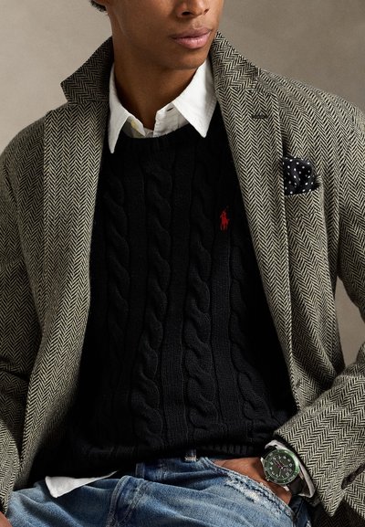 Šviesiai rudas ir juodas herringbone rašto švarkas dėvimas ant juodos kabelinės megztinio su raudonu logotipu. Po apačia balta marškinėliai, akcentuojami taškuoto kišeninio skarelės. Žalios spalvos laikrodis.