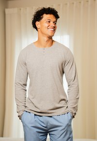 Jeune homme aux cheveux bouclés souriant, portant une chemise beige à manches longues et un pantalon rayé bleu, se tenant les mains dans les poches à l'intérieur.