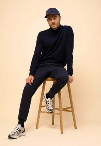 Námořnický žebrovaný svetr, tmavé kalhoty a bílé tenisky s černými akcenty. Model sedí na dřevěném stoličce na neutrálním pozadí.