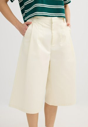 Persona que lleva pantalones cortos crema de tiro alto y pierna ancha con bolsillos, combinados con una camisa corta a rayas verdes y blancas, manos en los bolsillos.