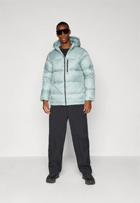 Άνδρας φορώντας ελαφρύ μπλε μπουφάν με κουκούλα, μαύρο cargo παντελόνι, μαύρα παπούτσια και υπερμεγέθη μαύρα γυαλιά ηλίου, στέκεται σε απλή φόντο.