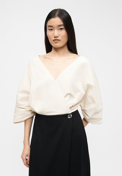 Γυναίκα με ίσια σκουρόχρωμα μαλλιά, ντυμένη με λευκό μπλουζάκι τύπου ντραπέ με V λαιμόκοψη και φαρδιές μανίκες, και μαύρη φούστα με κουμπιά στο πλάι.