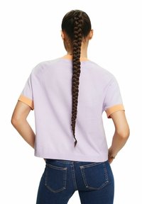 Fioletowy sweter z krótkim rękawem z kontrastowymi pomarańczowymi, prążkowanymi mankietami, noszony z ciemnoniebieskimi dżinsami. Z tyłu długie, splecione włosy.