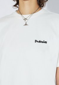 T-shirt en coton blanc à manches courtes, avec le texte brodé en noir "Drdenim" sur la poitrine. Deux colliers argentés avec pendentifs visibles.