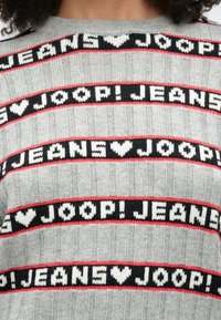 Γκρι πλεκτό πουλόβερ με οριζόντιες ζώνες κειμένου που γράφουν «JEANS ♥ JOOP!» σε λευκό χρώμα πάνω σε μαύρο και κόκκινο φόντο, με ριμπ υφή.