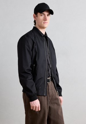 Junger Mann mit schwarzer Mütze, schwarzer Reißverschlussjacke, kariertem Hemd und brauner Hose, stehend vor einem schlichten hellgrauen Hintergrund.