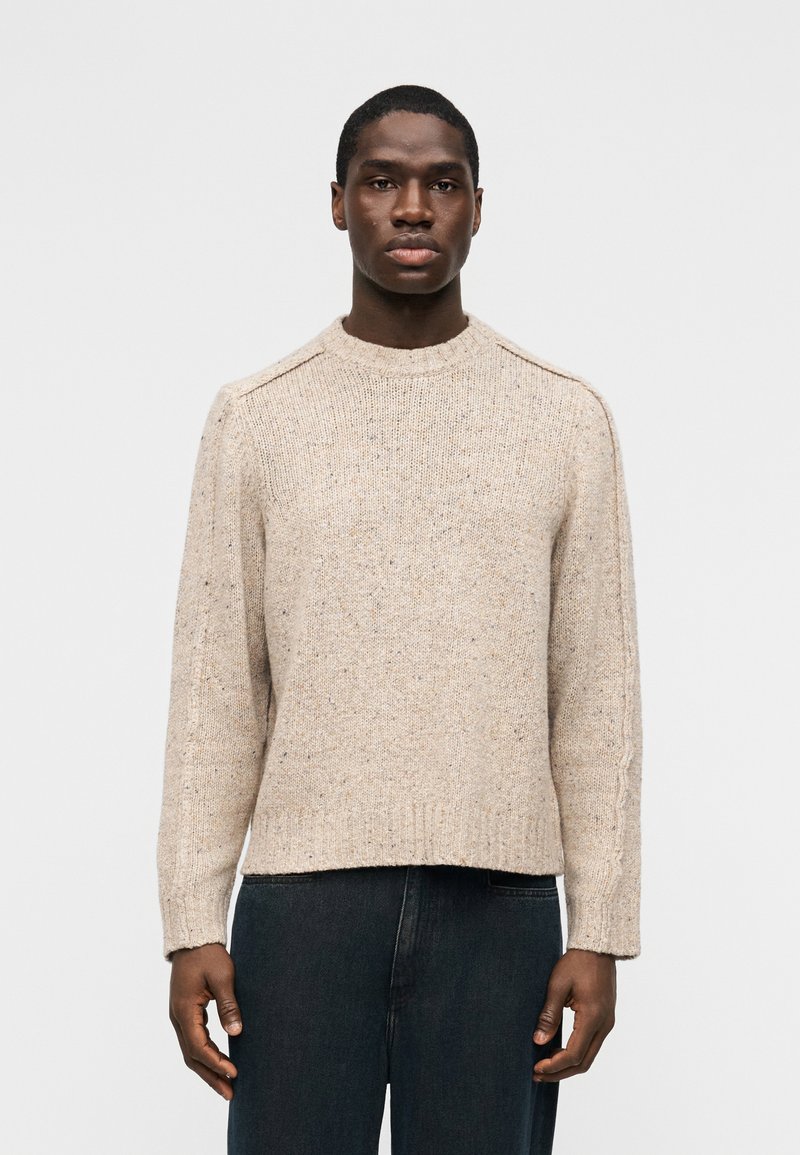 Άνδρας που φοράει μπεζ πλεκτό πουλ όβερ με στρογγυλή λαιμόκοψη και ριμπ μανσέτες, μαζί με σκούρο τζιν, στέκεται μπροστά σε απλό λευκό φόντο.