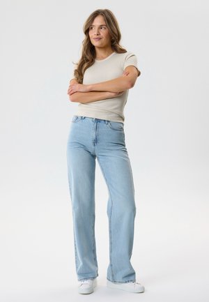 Žena s dlhými vlnitými vlasmi stojí so založenými rukami, oblečená v béžovom tričku s krátkym rukávom, svetlomodrých džínsoch a bielych teniskách.