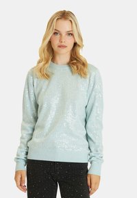 Jasnoniebieski sweter z cekinami pokrywającymi przód, prążkowanymi mankietami i dolnym wykończeniem, okrągłym dekoltem oraz miękką fakturą. W połączeniu z ciemnymi dżinsami.