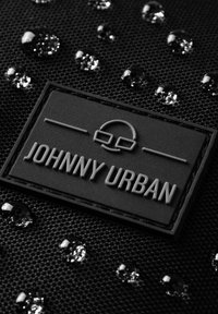 Tessuto nero testurizzato resistente all'acqua con una patch in gomma con la scritta "JOHNNY URBAN". Sono presenti motivi circolari e chiare gocce d'acqua.