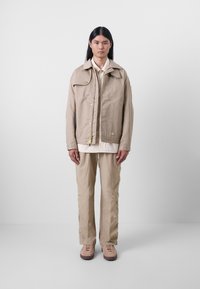 Veste beige à poches multiples avec un col, fermeture par bouton et cordon de serrage. Associée à un pantalon beige et des chaussures à lacets brun clair. Texture lisse.