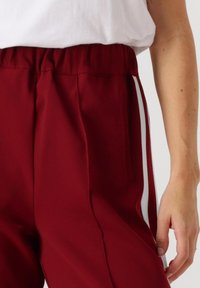 Bordeaux atletische broek met een witte zijstrook, elastische tailleband en zakken; gladde textuur met een relaxte pasvorm.