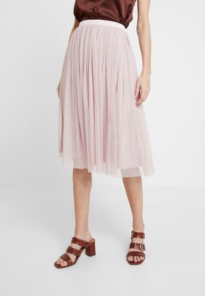 Vrouw met een lichtroze geplooide midi-rok, een bruine mouwloze blouse en bruine hakken met dubbele bandjes.