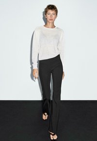 Hellgrauer Pullover mit langen Ärmeln und weicher Textur, kombiniert mit schwarzen, enganliegenden Hosen. Das Modell trägt braune Sandalen und steht vor einem dunklen Hintergrund.