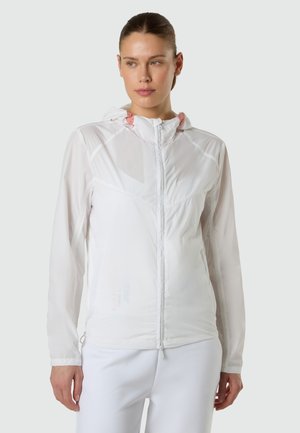 Mujer joven con cabello oscuro recogido, viste una chaqueta blanca ligera con cremallera y capucha, y pantalones blancos a juego, mirando ligeramente hacia un lado.