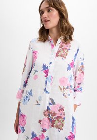 Witte linnen shirtjurk met een roze, blauwe en paarse bloemenpatroon, knopenlijst en driekwartmouwen. Zachte textuur en losse pasvorm.