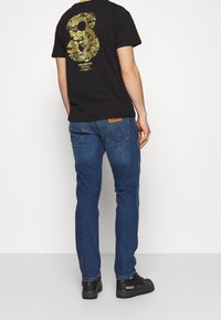 Svart t-shirt med guldtryck på baksidan, ihop med mörkblå denimjeans och svarta sneakers. Enkelt, figurnära design och avslappnad stil.