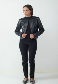 Czarna skórzana kurtka z okrągłym dekoltem i zapięciem na zamek z przodu, stylizowana z czarnymi dopasowanymi spodniami i płaskimi butami. Gładka faktura i dopasowany krój.