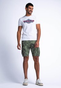 Camiseta blanca de algodón con gráfico en rojo y azul que dice "PETROLIND", combinada con pantalones cortos de camuflaje verde con bolsillos laterales. Incluye calzado casual.