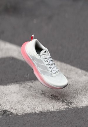 Zapatilla de correr blanca y rosa con cordones, flotando sobre una superficie de asfalto gris con marcas blancas y pequeñas partículas de grava.