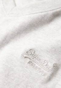 Lysegrå bomuldsstof med en blød tekstur; har en V-hals-design og broderet "SuperDry" logo i hvid tråd.