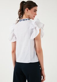 Balta lininė viršutinė dalis su trumpomis ruffles rankovėmis, išsiuvinėtas tamsiai mėlynas floros raštas prie iškirpčių, ir laisvas siluetas. Pateikiamas galinis vaizdas.