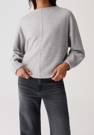 Kobieta ubrana w jasnoszary sweter z długim rękawem z przeszyciem na środku oraz ciemnoszare dżinsy, ręce spoczywają w okolicy talii.