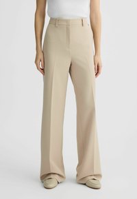 Pantaloni beige a gamba larga con pieghe frontali, tessuto morbido, vita alta e tasche laterali, abbinati a scarpe chiare.
