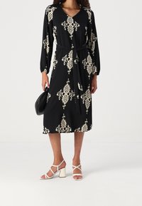 Vestido negro con patrón de paisley crema, mangas largas, escote en V, cintura ceñida con cinturón y largo midi. Combinado con sandalias blancas de tacón bloque con tiras.