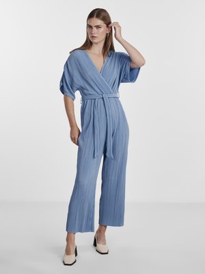 Salopette plissée bleu clair avec un décolleté en V profond, des manches larges et une taille ceinturée, associée à des chaussures à talons carrés crème.