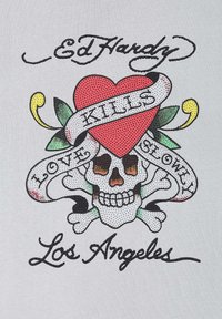 Graphic t-shirt featuring a skull with a heart, surrounded by leaves and ribbons. Colors include red, black, green, and yellow on a light gray background.