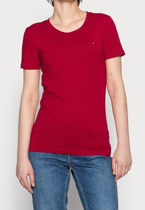 Person trägt ein eng anliegendes rotes T-Shirt mit kurzen Ärmeln und blaue Jeans, mit einem kleinen Logo auf der linken Brust.