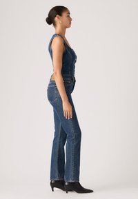 Γυναίκα σε πλαϊνή όψη, ντυμένη με αμάνικη ολόσωμη φόρμα από τζιν και μαύρα μποτάκια με μυτερή μύτη, μπροστά σε απλό φόντο.