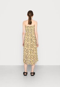 Žluté šaty bez ramínek s květinovým vzorem, s nařaseným korzetem a midi délkou, doplněné černými sandály. Texturovaná a lehká tkanina.