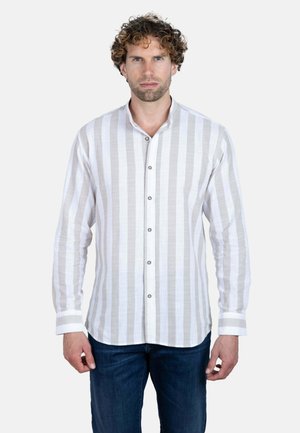 Hombre con cabello rizado que lleva una camisa de manga larga blanca y gris claro con rayas verticales y vaqueros azul oscuro, de pie, mirando al frente.