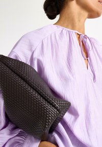 Blusa viola con dettaglio al collo arricciato, abbinata a una borsa in pelle intrecciata scura con forma arrotondata e superficie testurizzata.