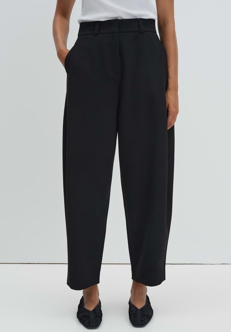 Woman wearing black high-waist wide-leg trousers with side pockets and black pointed flats against a plain light background.