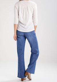 Weißes langärmliges Oberteil mit Faltendetail auf der Rückseite, kombiniert mit blauen weit geschnittenen Jeans und hautfarbenen High Heels. Glatte Textur überall.