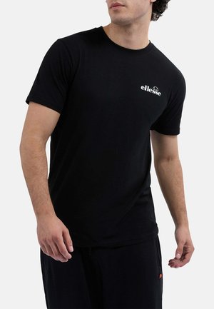 Mann in einem schwarzen ellesse T-Shirt und schwarzen Hosen, der vor einem schlichten weißen Hintergrund steht und leicht zur Seite schaut.